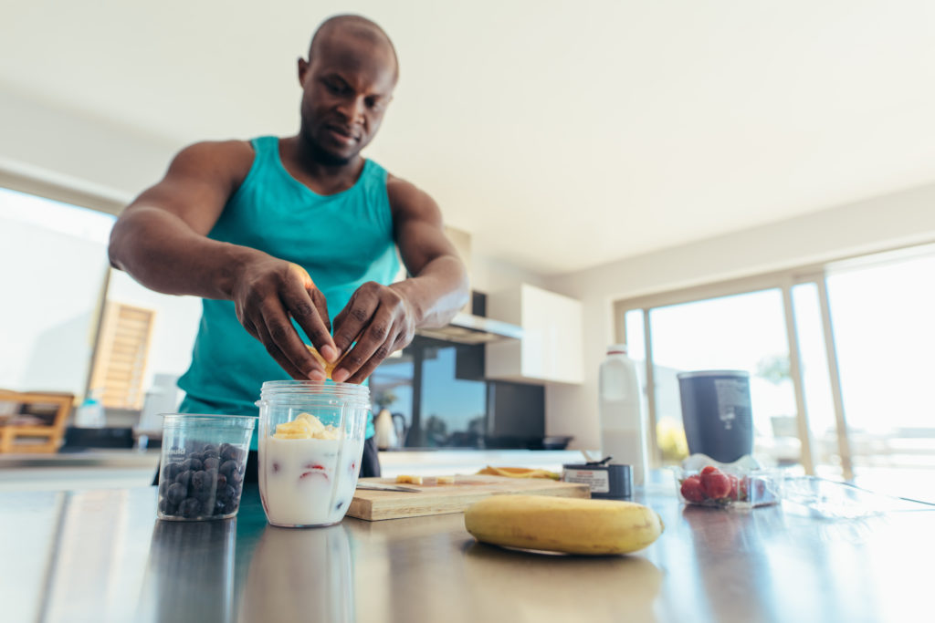 Man preparing milk shake in kitchen. Athletic man adding fruits to jar of milk.